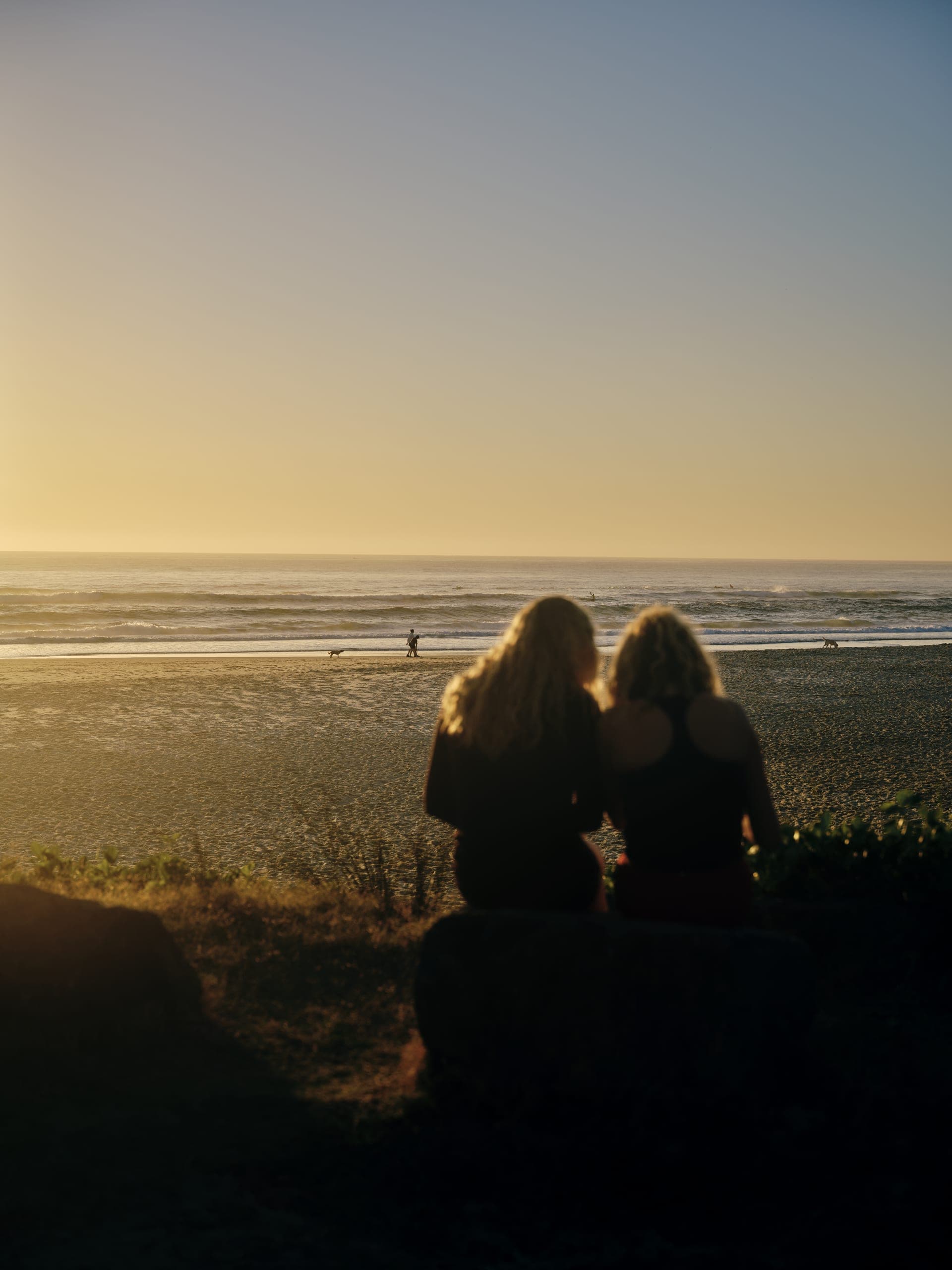 Two people sitting side by side above the beach, watching the surf at sunset